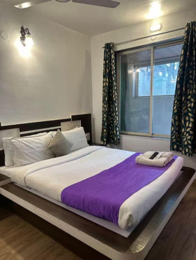 A hotel room with a bed, a window with curtains, a ceiling fan, and wall-mounted lights.