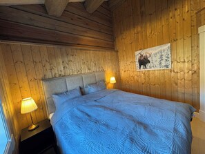 A room with wooden walls, a bed with a blue quilt, two bedside lamps, and a framed picture of a moose. - Familycabin at a lake (Ringsaker)