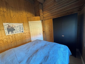 Une pièce aux murs en bois, un lit recouvert d’une couvre-lit à motifs, une porte et une armoire de couleur sombre.