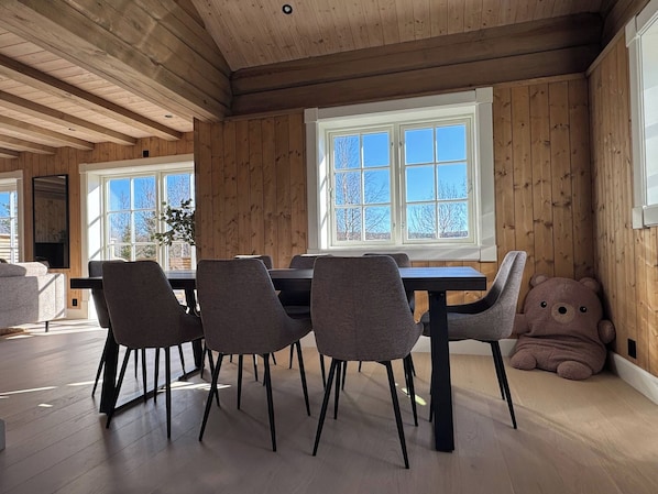 Un coin repas avec un décor intérieur en bois, une grande fenêtre et un ours en peluche posé sur le sol.