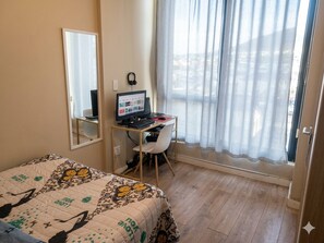 Ein Schlafzimmer mit einem Bett, einem Schreibtisch mit Computer, einem Stuhl und einem Fenster mit Vorhängen.
