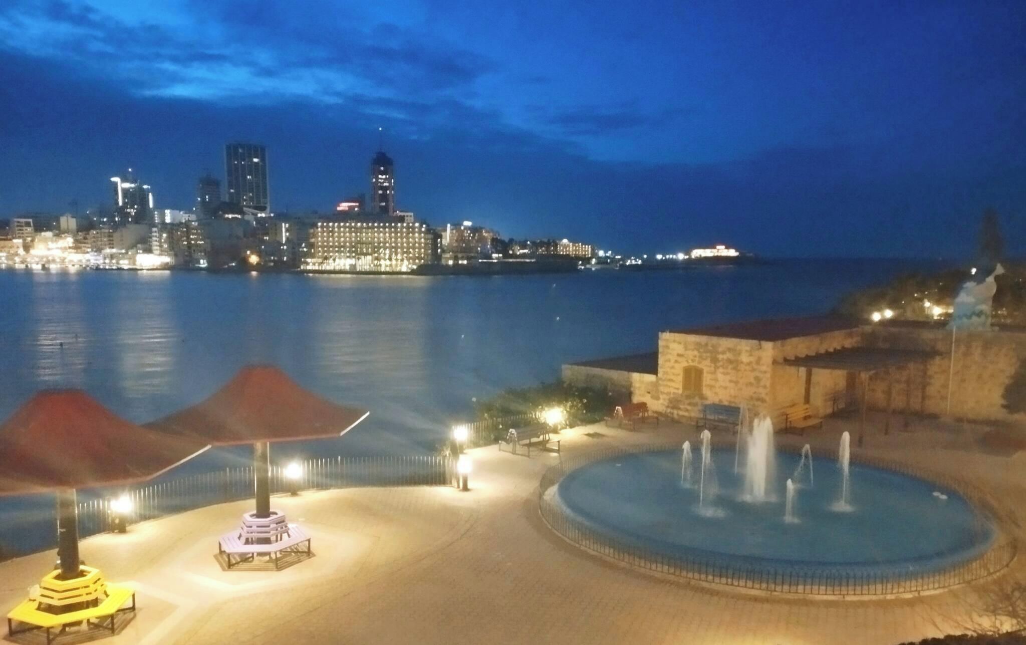 A waterfront promenade with a fountain, a covered seating area, and a view of a city skyline at dusk.