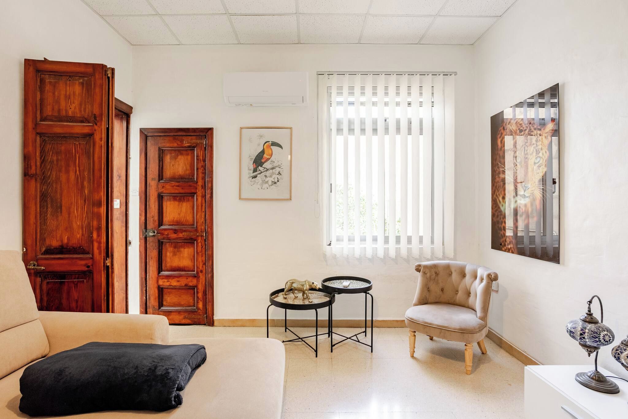 Una sala de estar con un sofá, un sillón, dos mesitas, un ventanal con persianas y un aire acondicionado instalado en la pared.
