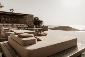 Ein sonnendurchfluteter Außenbereich mit gepolsterten Sofas und einem Tisch, der mit Speisen und Getränken gedeckt ist, mit Blick auf ein Gewässer.