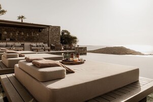 Un espacio al aire libre iluminado por el sol con sofás acolchados y una mesa dispuesta con comida y bebidas, con vista a un cuerpo de agua.