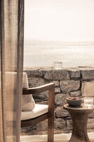 Un muro de piedra con vista al mar, una silla de madera con cojín, una mesita redonda con un cuenco y vasos.