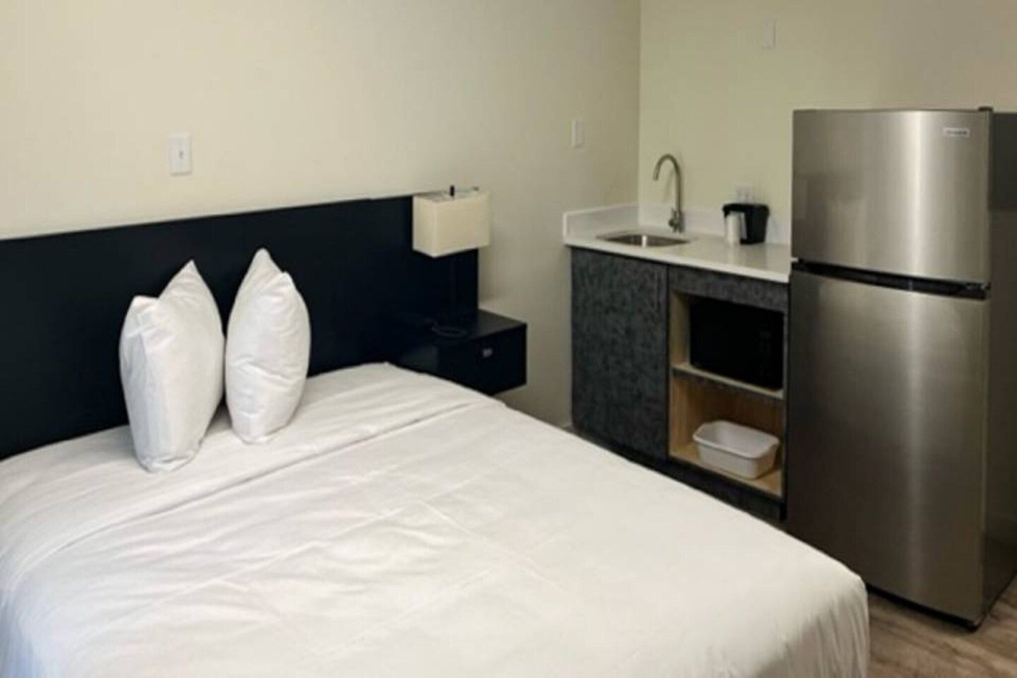 A hotel room with a bed, a nightstand, a lamp, a sink, and a refrigerator.