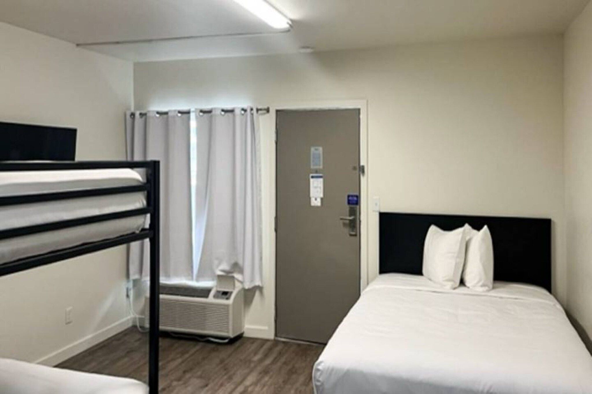 A hotel room with a bunk bed, a single bed, a door with a window, and a wall-mounted air conditioning unit.