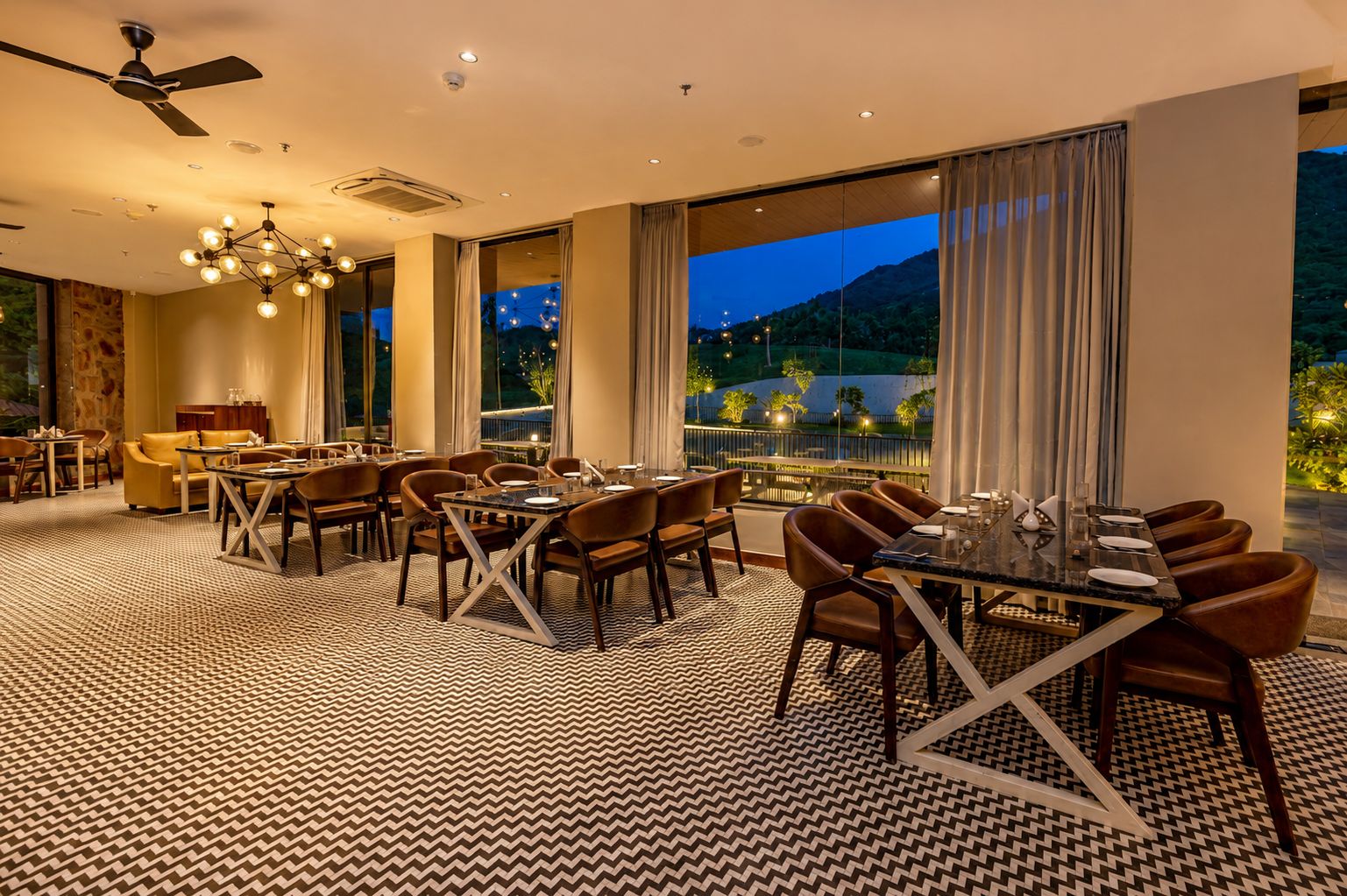 Un comedor de hotel con una gran mesa dispuesta para una comida, rodeada de sillas, y vistas del exterior al anochecer.