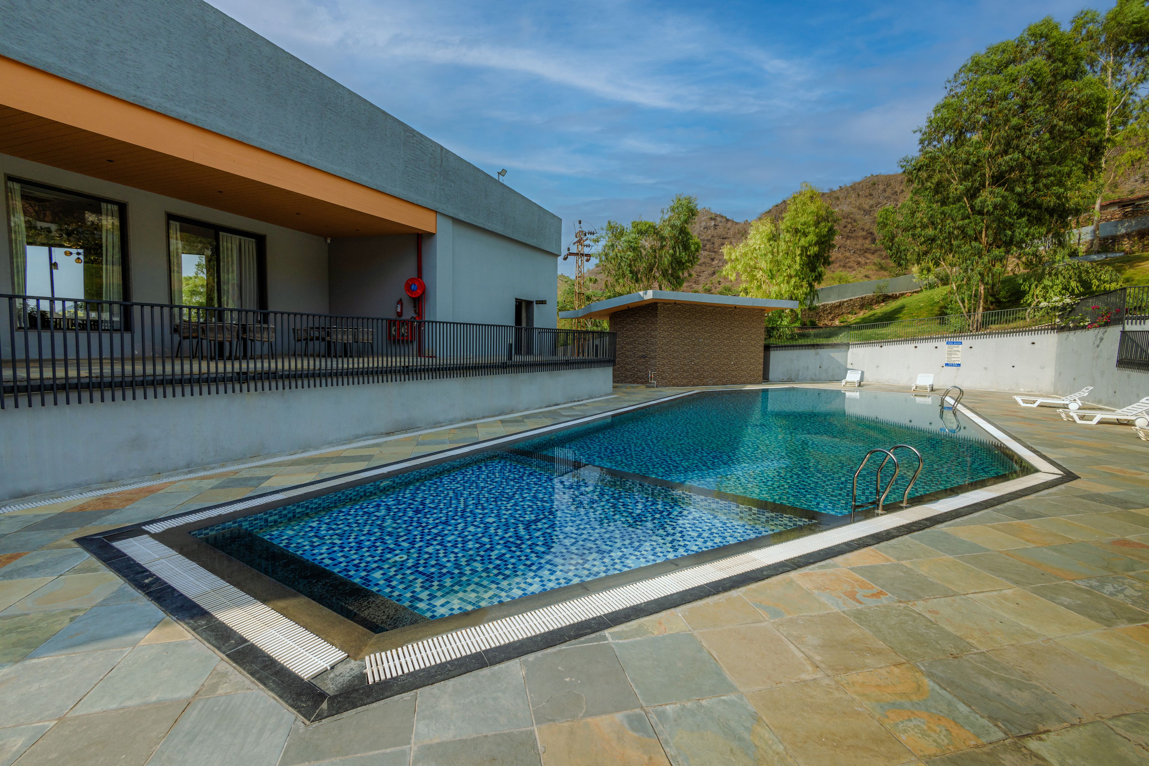 Una piscina con solárium de baldosas, una estructura pequeña junto a la piscina y un edificio con grandes ventanales.