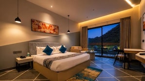 A hotel room with a bed, bedside table, lamp, artwork, and a view of the outdoors. - Ullasa by Aranyam  Luxury Resort Udaipur (udiapur)