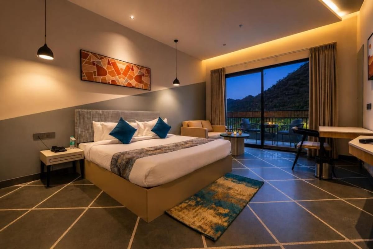 Habitación de hotel con cama, mesita de noche, lámpara, obra de arte y vistas al exterior.