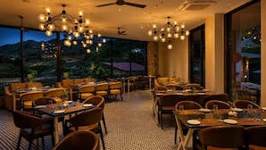 A restaurant with tables and chairs arranged for dining, large windows offering a view of the outdoors, and a ceiling fan. - Ullasa by Aranyam  Luxury Resort Udaipur (udiapur)