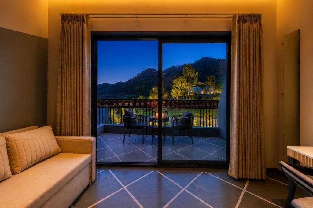 A room with a sofa, a table, and a balcony with a view of a mountain and trees.