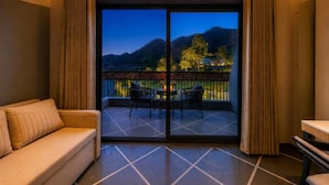 A room with a sofa, a table, and a balcony with a view of a mountainous landscape. - Ullasa by Aranyam  Luxury Resort Udaipur (udiapur)