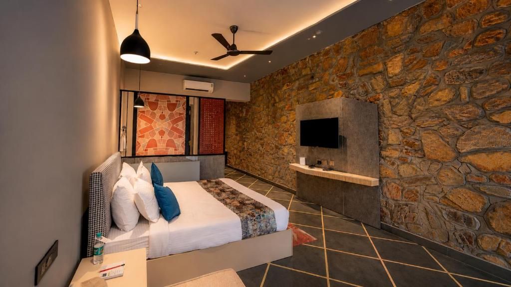 A modern bedroom with a stone wall, a bed with pillows, a bedside table, a flat-screen TV, and a ceiling fan.