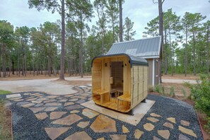 A wooden sauna structure with a sloped roof, situated on a stone-tiled patio surrounded by a forest. - Piney Woods Retreat w/ Sauna, Fire Pit & Games (Willis)