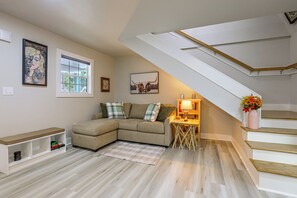 Ein modernes Wohnzimmer mit einer Eckcouch, einem kleinen Tisch mit einer Lampe, einer Treppe und Kunstwerken an den Wänden.