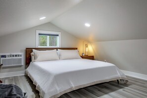 A bedroom with a sloped ceiling, a bed with white bedding, a window with a view of trees, a wall-mounted air conditioner, and a bedside lamp. - Wooded Piney Woods Tiny Home w/ King Loft & Deck (Willis)