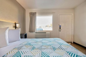 A hotel room with a bed, a window with curtains, a door, and a wall-mounted air conditioning unit. - 1 King Bed | Micfridge (Albuquerque NM)