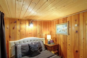 Ein Zimmer mit Holzwänden, einem Bett, einem Nachttisch, einer Lampe und einem Gemälde an der Wand.