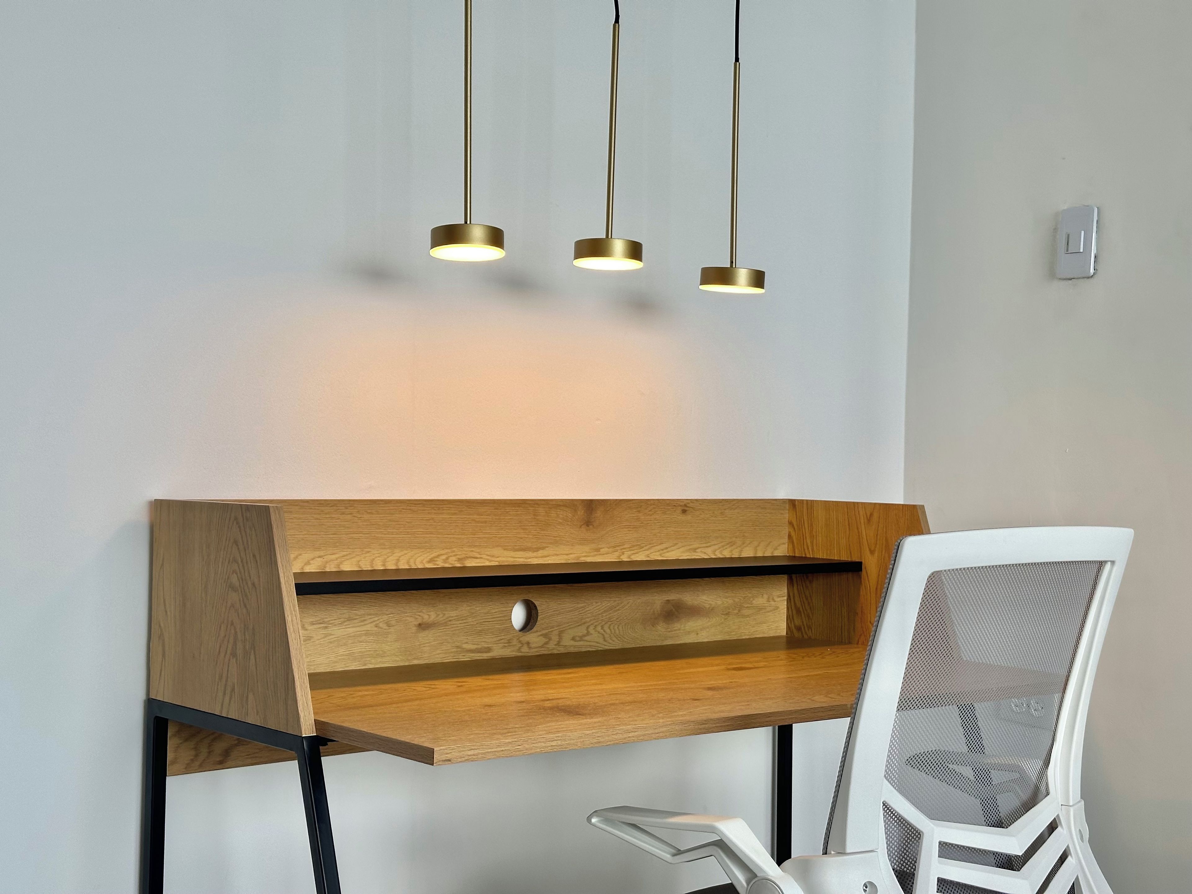 Un bureau en bois, une chaise à dossier en treillis métallique, un tiroir et trois lampes suspendues.