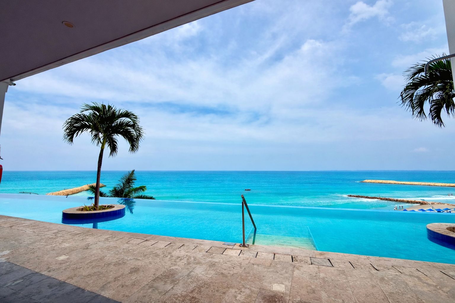 Vista a la piscina con cielo azul despejado, playa de arena y palmeras.