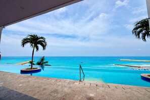 Vista a la piscina con cielo azul despejado, playa de arena y palmeras.