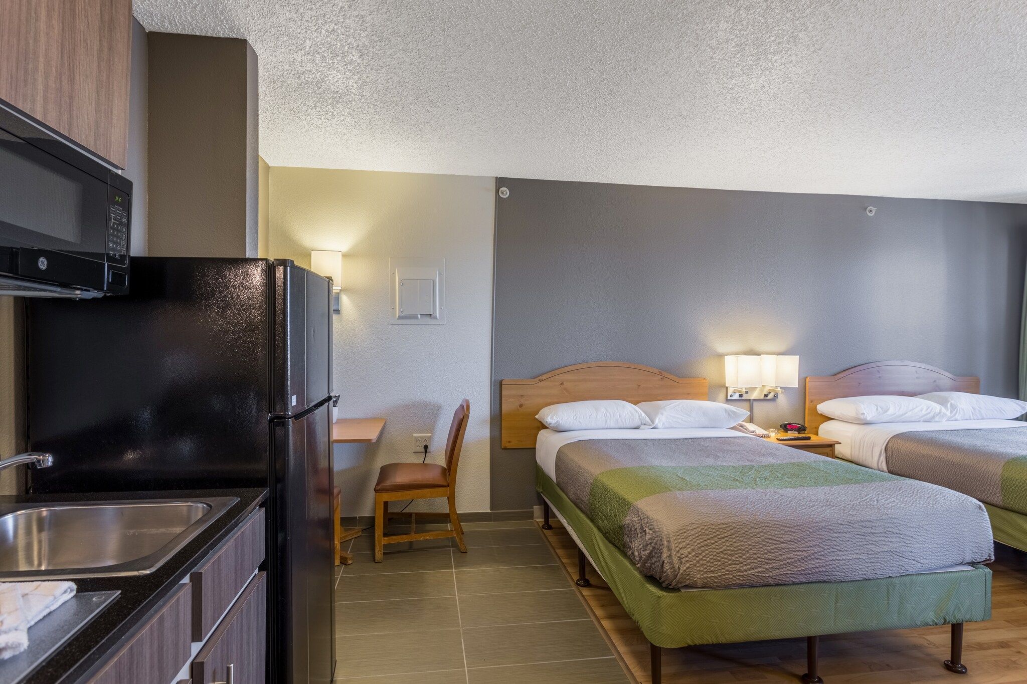 A hotel room with a kitchenette, a single bed, a second bed, a chair, and a nightstand with a lamp.