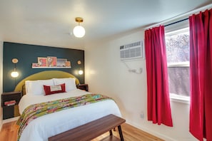 Ein Schlafzimmer mit einem Bett, Nachttischen, einer Bank, einer Wand-Klimaanlage und einem Fenster mit roten Vorhängen.