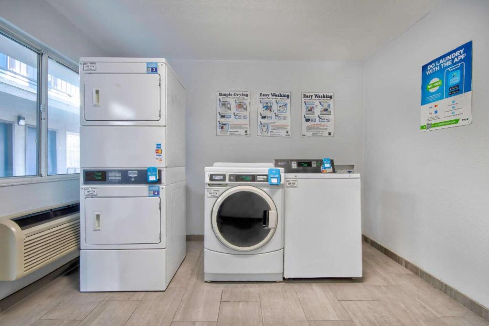 A laundry room with a dryer, washer, and dryer with a control panel. There are posters on the wall advertising laundry services.