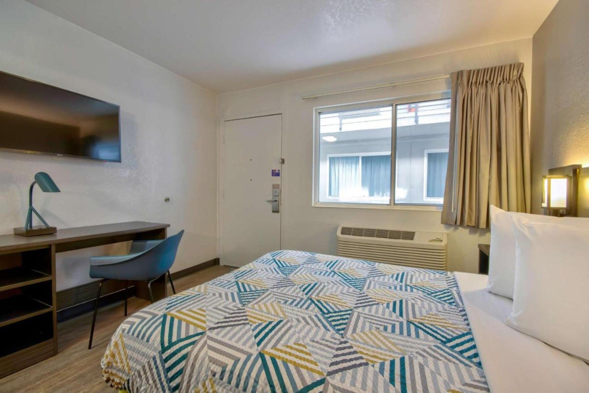 Ein Hotelzimmer mit Bett, Schreibtisch, Stuhl, einem Fenster mit Vorhängen und einer Wandlampe.