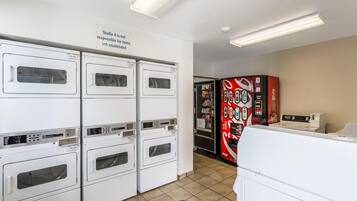 A laundry facility with multiple washing machines and dryers, a Coca-Cola vending machine, and a sign indicating Studio 6 is not responsible for unattended items.