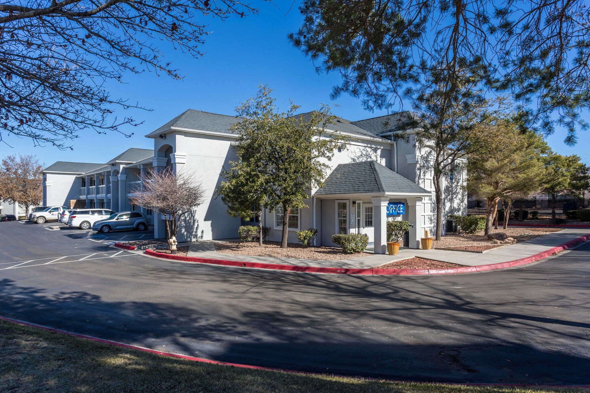 A building with a sign indicating an office, a parking lot with several cars, and a red curb along the street.