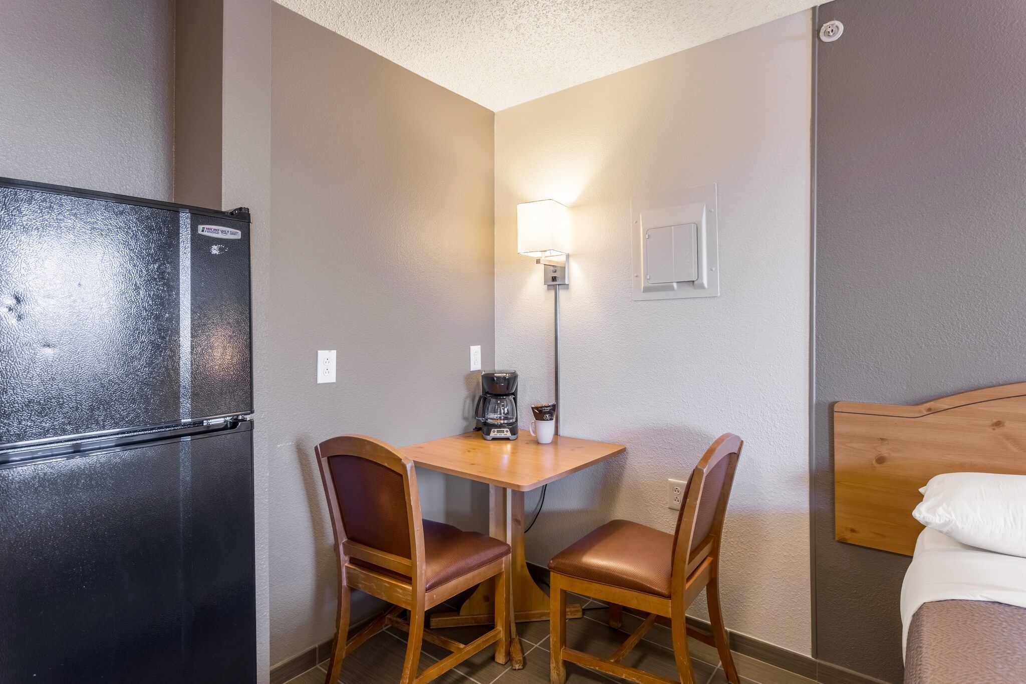 A compact hotel room with a kitchenette, a small table with two chairs, a bed, and a wall-mounted light fixture.