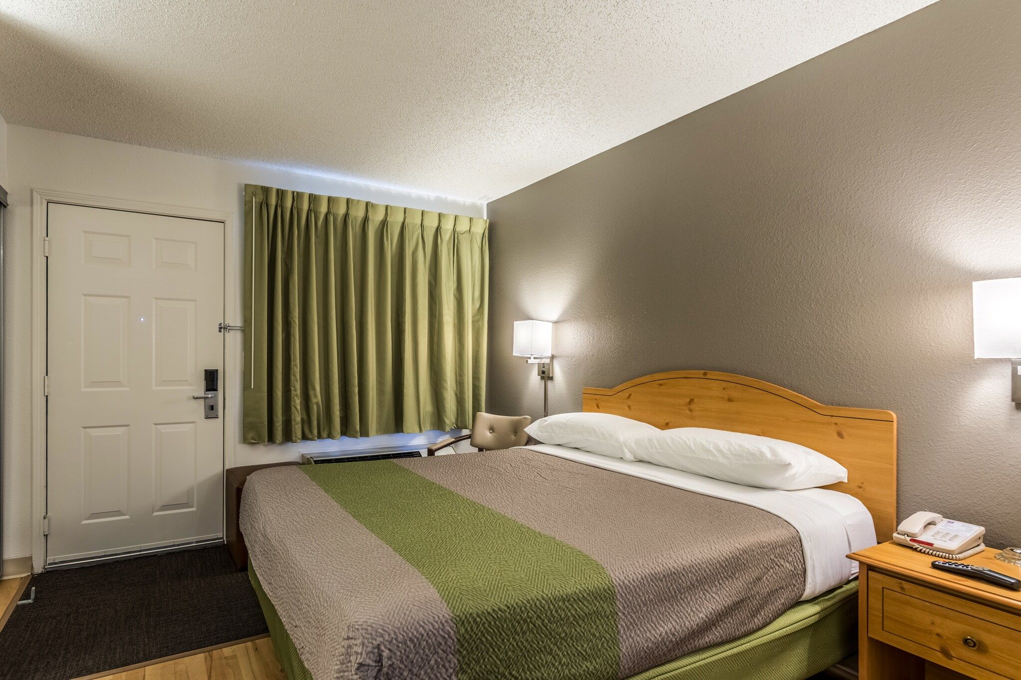 A hotel room with a bed, a bedside table with a phone, a lamp, and a door.