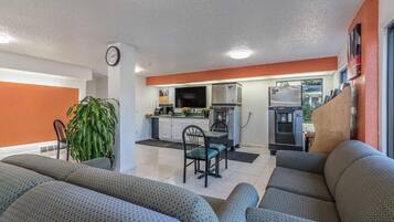 A modern living room with a sofa set, a dining area with chairs, a kitchen with appliances, and a clock on the wall.