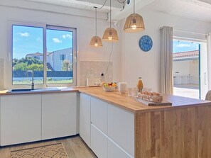 A modern kitchen with a wooden island, pendant lights, a clock, and a view of the outside. - Les Salines by Interhome (Ile d'Oléron)