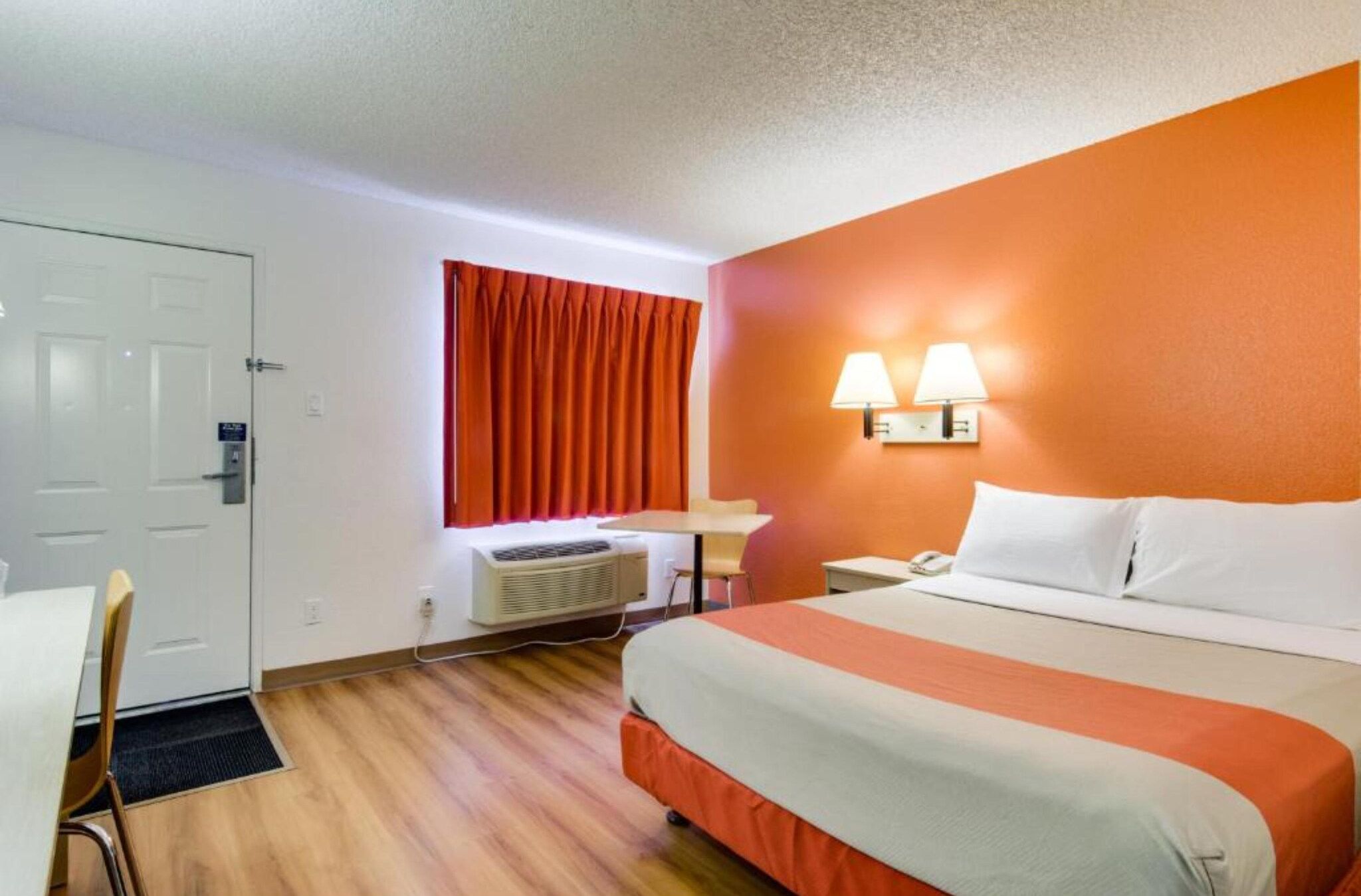 Ein Hotelzimmer mit Bett, Schreibtisch, Stuhl, einem Fenster mit Vorhängen und einer Wandlampe.