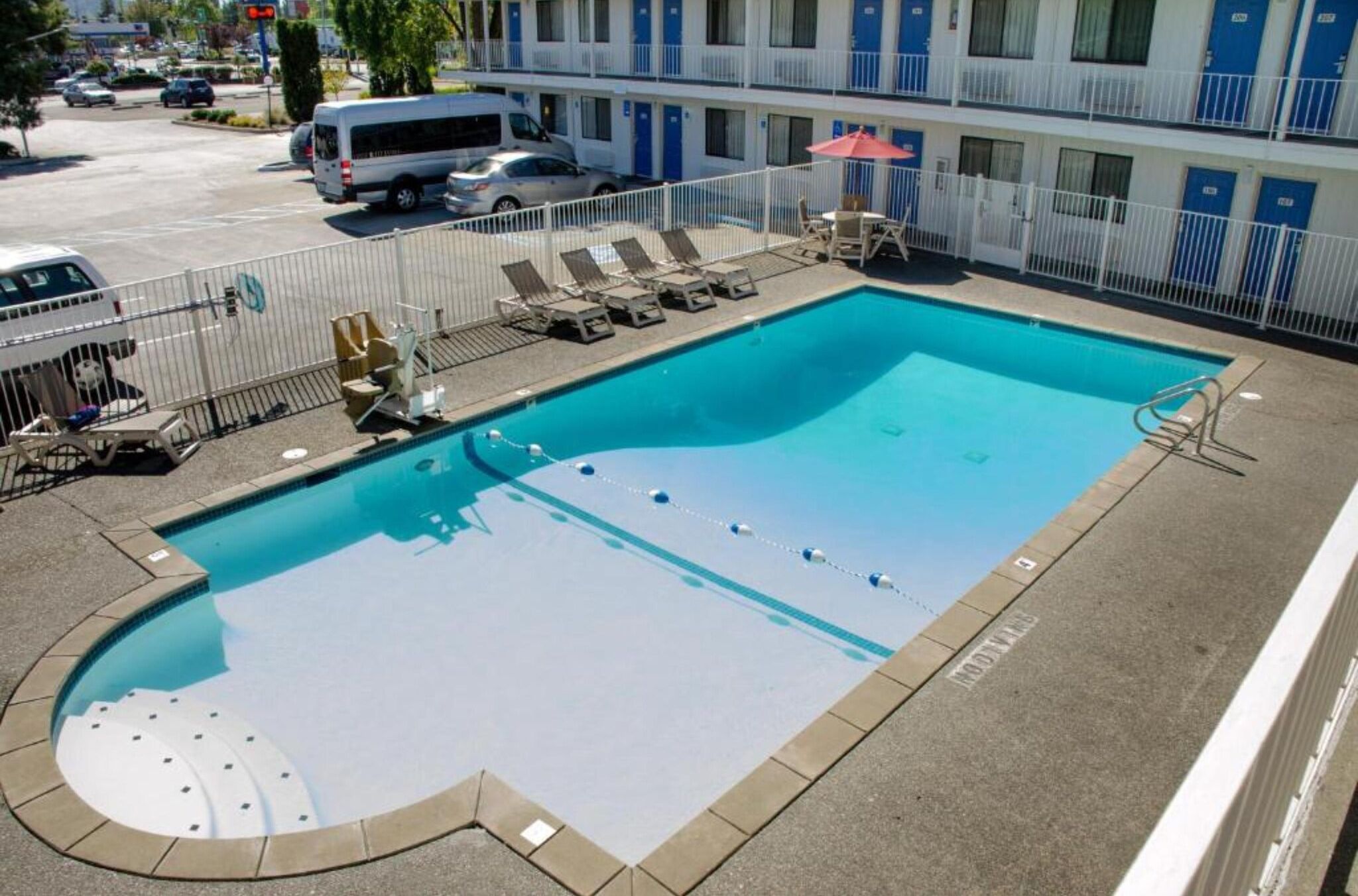 A swimming pool with lounge chairs and a sun umbrella, surrounded by a fence, with a building in the background.