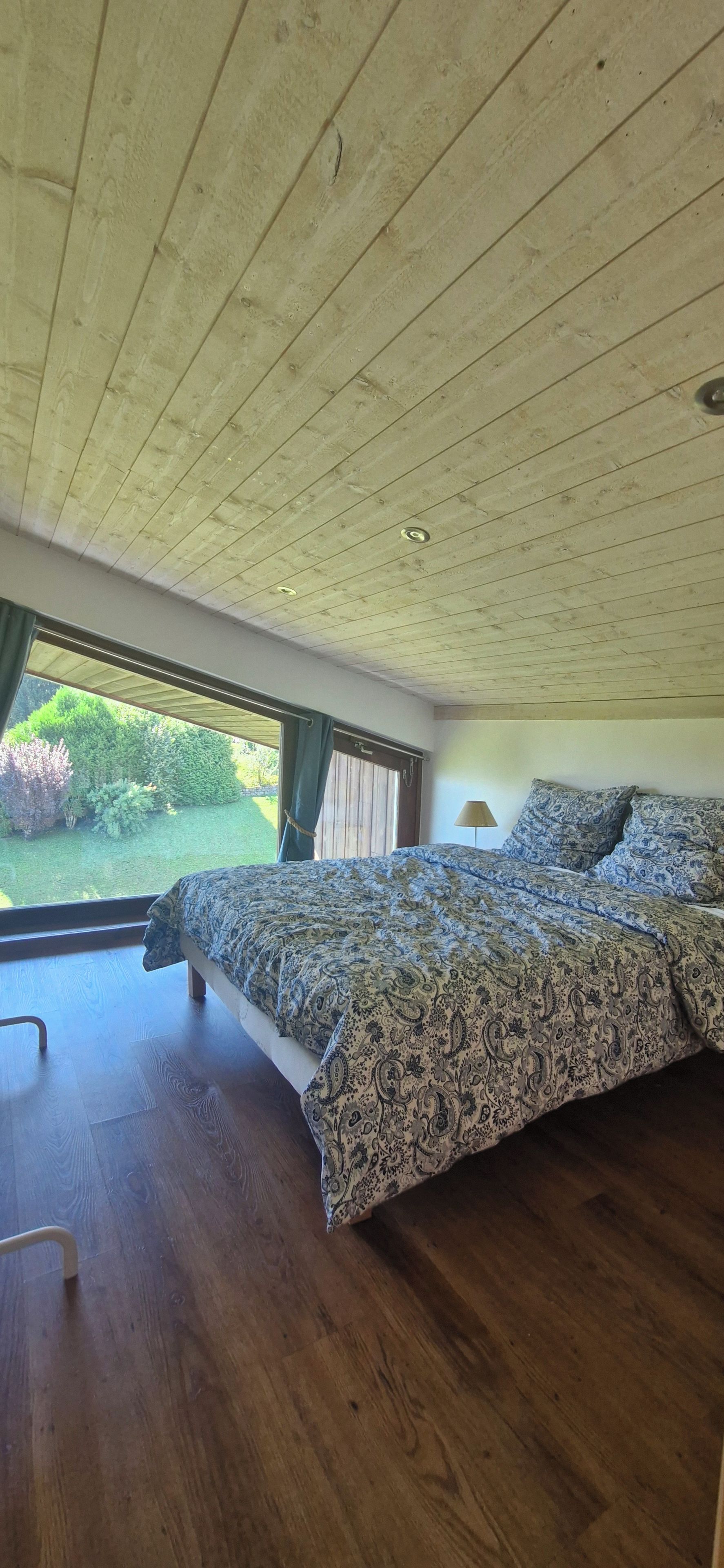 Un dormitorio con techo de madera, una cama grande con ropa de cama estampada, vista a zonas verdes y suelo de madera.