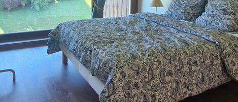 Un dormitorio con techo de madera, una cama grande con ropa de cama estampada, vista a zonas verdes y suelo de madera.