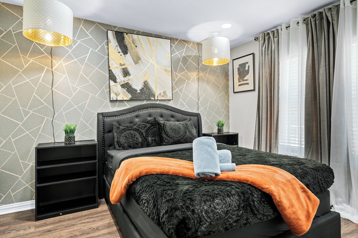 Ein modernes Schlafzimmer mit einem gesteppten Kopfteil, einem großen abstrakten Gemälde, zwei Pendelleuchten, einem Bett mit dunkler Decke und orangem Überwurf sowie einem Nachttisch mit Pflanze.