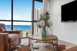 A modern living room with a leather sofa, a round coffee table, a vase with flowers, a remote control, and a large window offering a view of the ocean and high-rise buildings. - 5 minute walk to pacific fair and beach! 2BR prime location high rise apartment (Mermaid Beach)