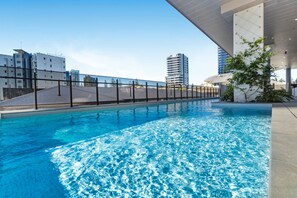 Une piscine à l’eau d’un bleu limpide, entourée d’une clôture et bordée de bâtiments en arrière-plan.