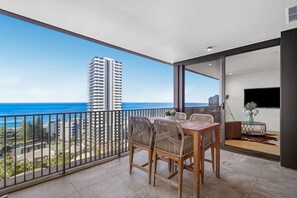 A balcony with a dining table and chairs, a TV, and a view of the ocean and a tall building. - 5 minute walk to pacific fair and beach! 2BR prime location high rise apartment (Mermaid Beach)