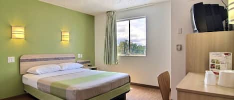 A hotel room with a bed, a desk with a chair, a window with a view, and a wall-mounted air conditioning unit.