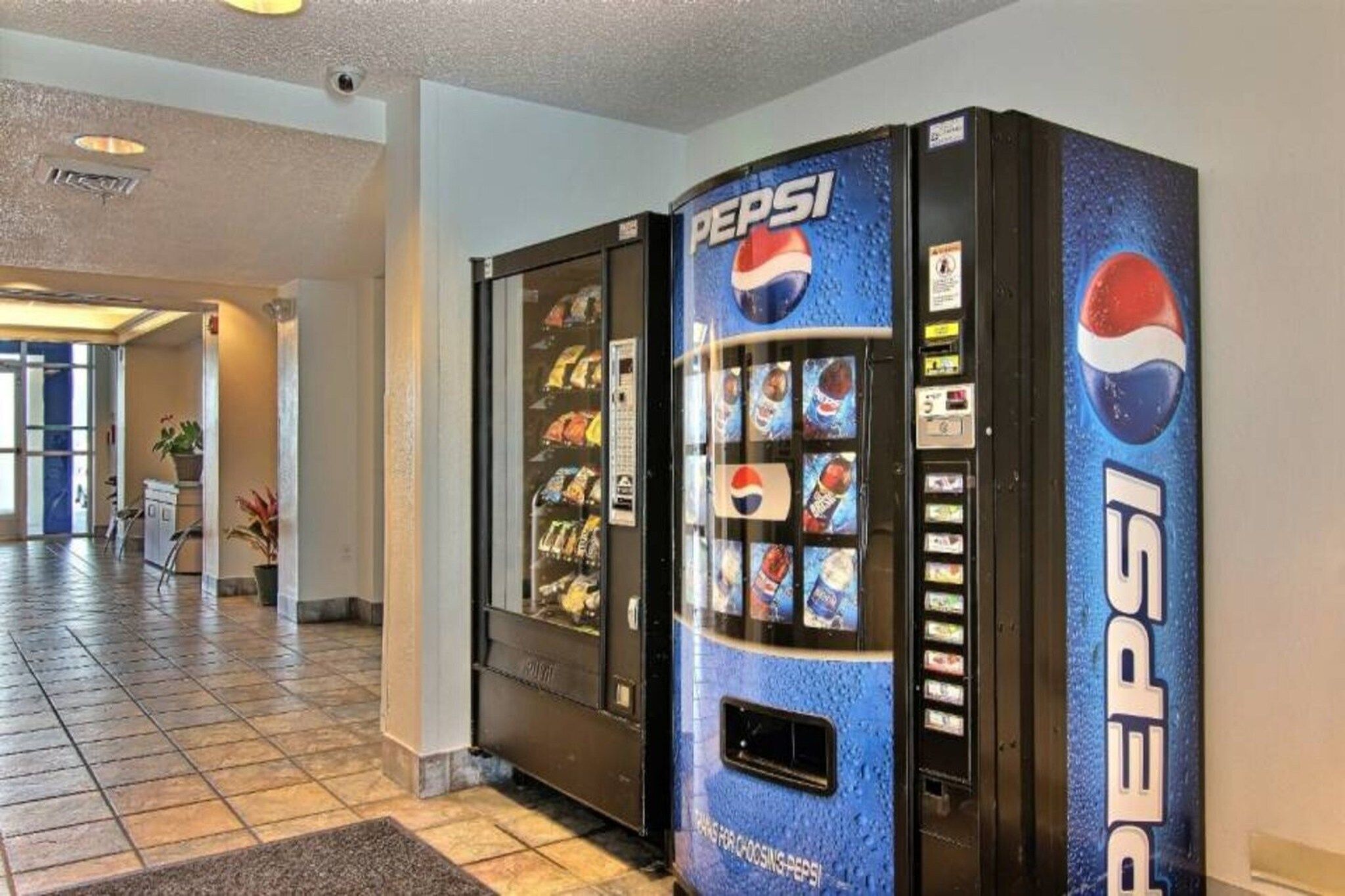 A Pepsi vending machine in a hotel lobby with a tiled floor and a plant in the background.