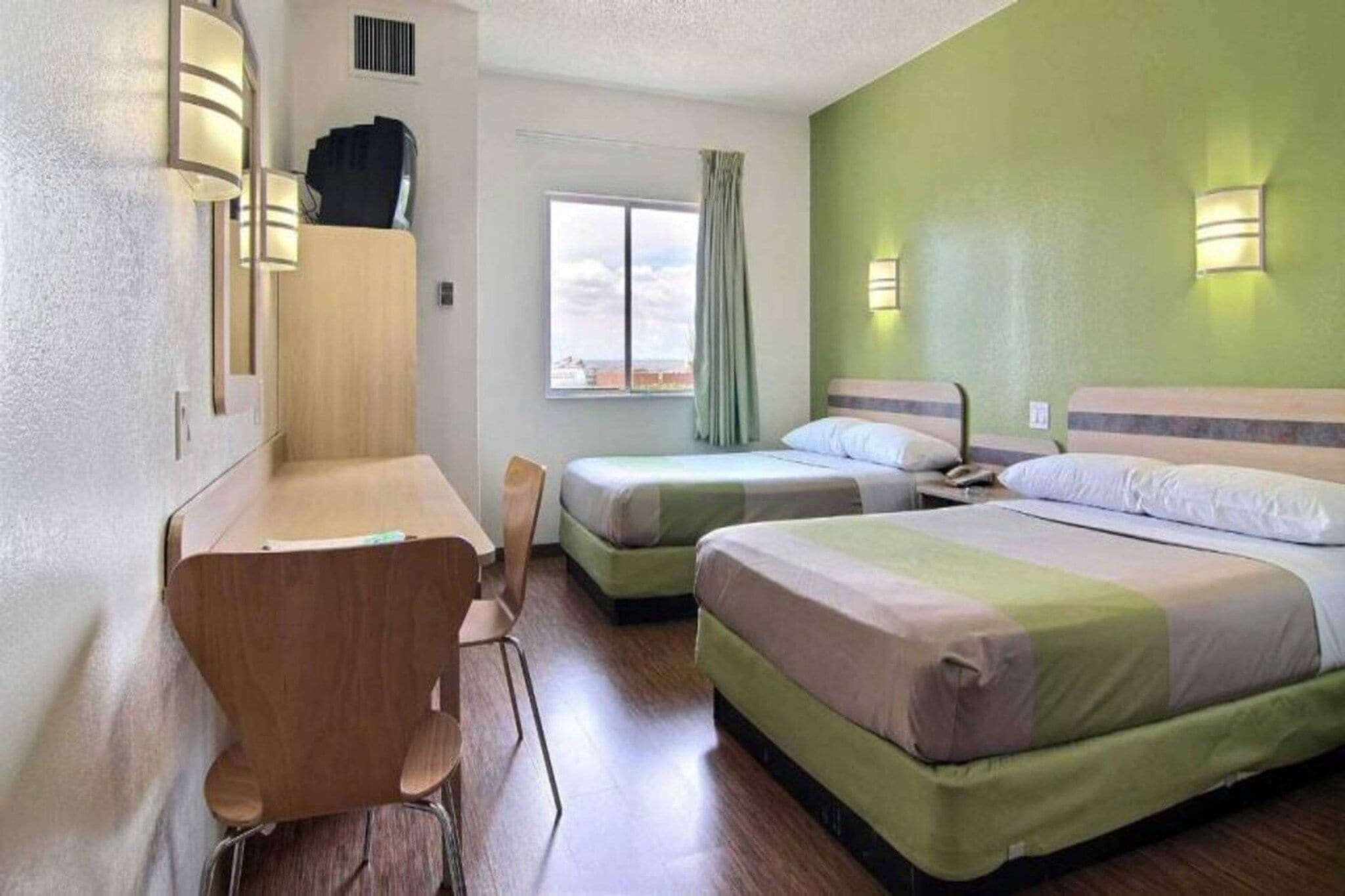 A hotel room with a desk, two single beds, a chair, and a window with curtains.