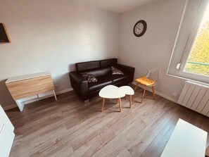 Un salon moderne avec un canapé en cuir noir, une petite table d’appoint blanche et une petite chaise en bois. Une horloge murale est fixée au mur.